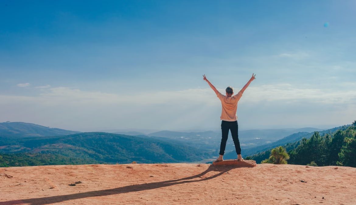A man raises his arms in triumph on a rocky mountain summit overlooking a vast landscape